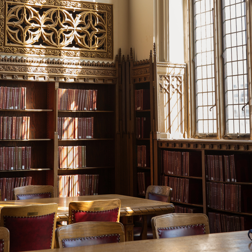 The Great Reading Room with wooden tables and chairs in the forefront. In the background are bookshelves, multi-pane windows, and ornate decorative wall architecture.